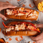Mano che solleva un lobster roll Mason's sopra un vassoio di patatine